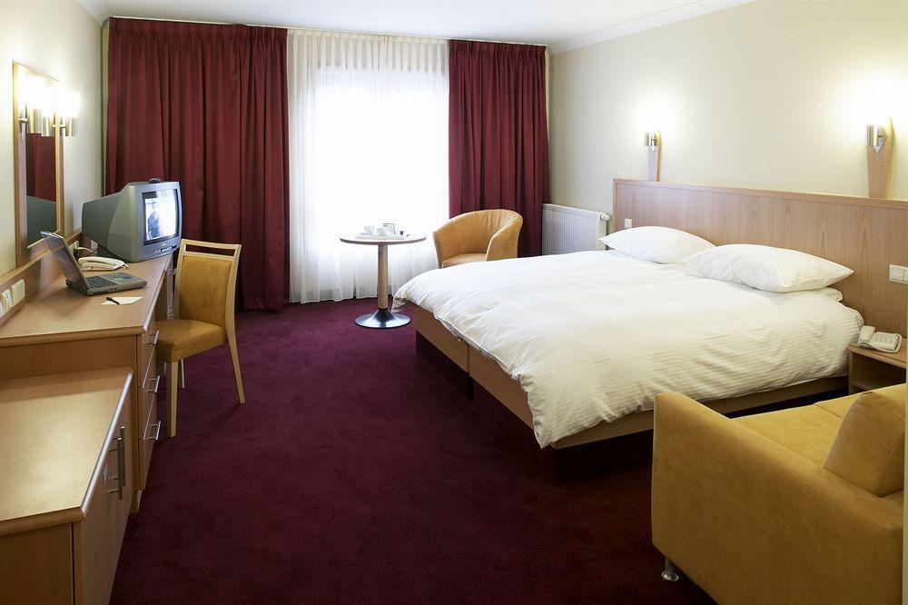 Clayton Hotel Dublin Airport Coolock Camera foto The photo shows a hotel room featuring a double bed with white linens and pillows. There are two bedside lamps mounted on the wall above the bed. To one side, there's a small round table with two chairs. Opposite the bed, a wooden desk with a chair i